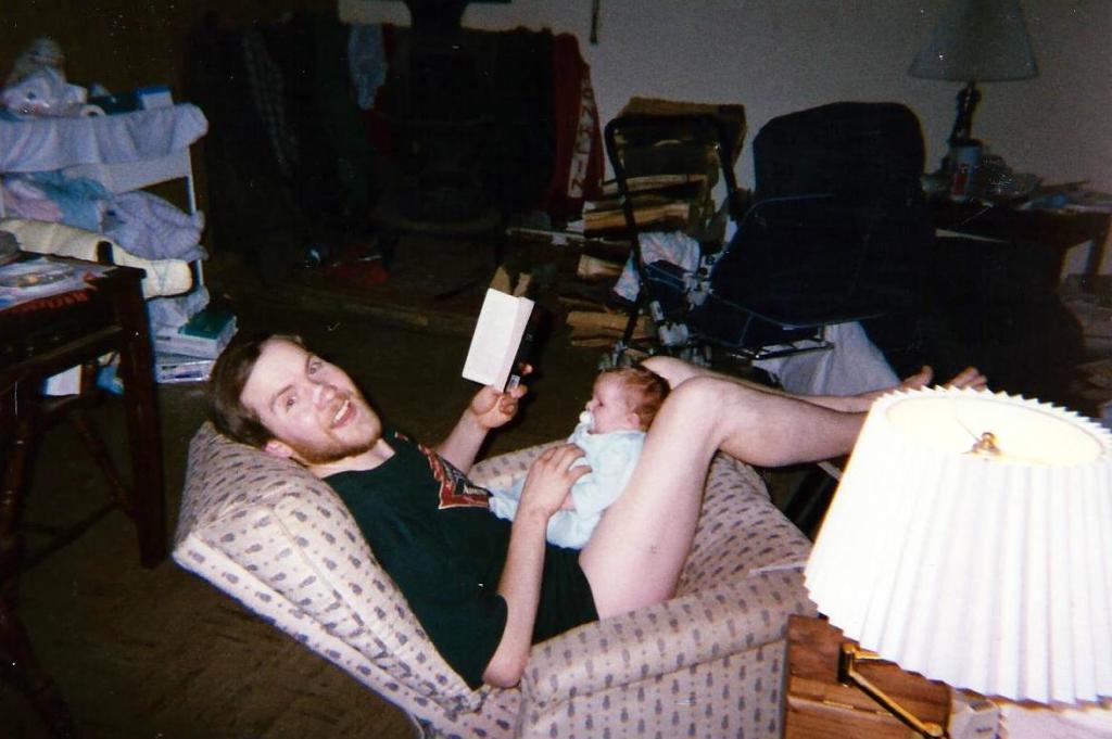 Reading while holding Carly. A lot of clutter of stuff,  along with a lot of firewood near the stove.